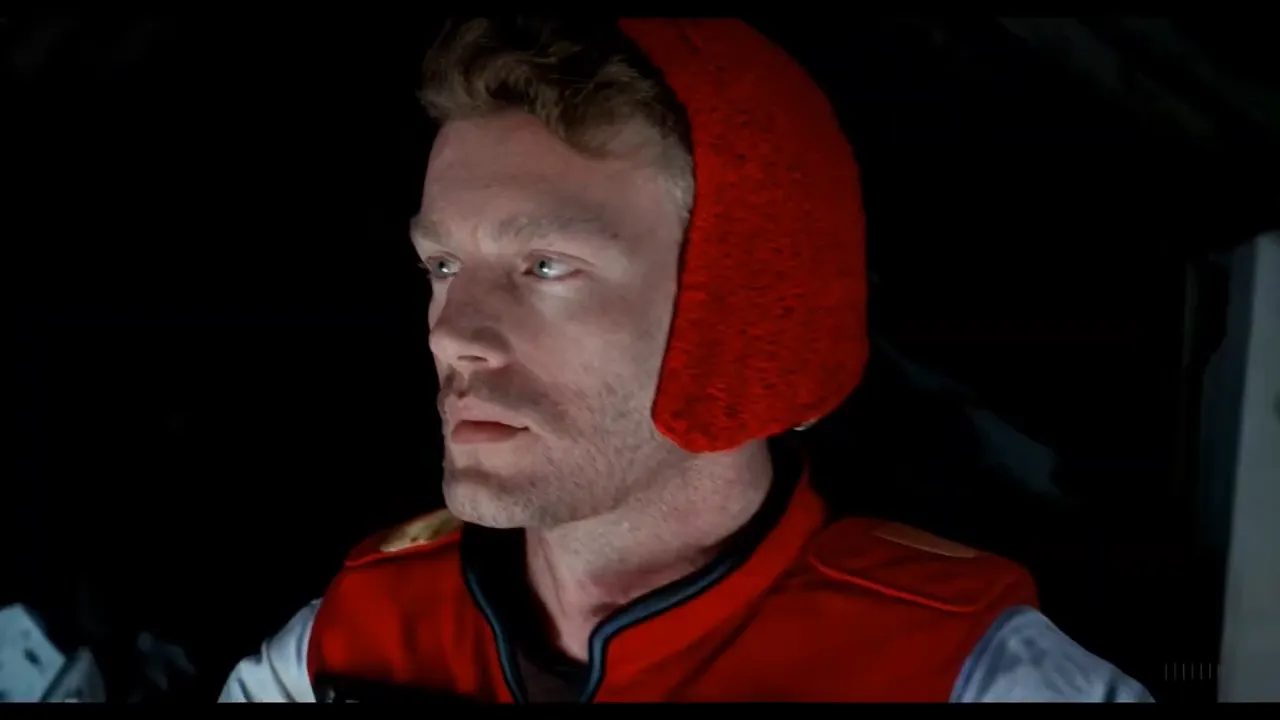 A man in a red knitted hat and red space suit looking to one side inside a dim spacecraft cabin.